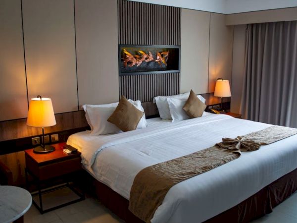 A modern hotel room with a neatly made bed, pillows, bedside lamps, and a wall fireplace with burning logs.