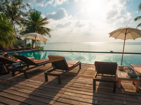A tropical oceanfront resort deck with lounge chairs, umbrellas, a swimming pool, and calm sea under a cloudy sunset sky.