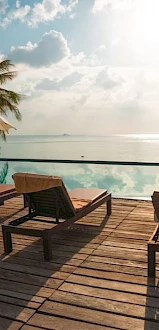 A seaside resort deck with pool, lounge chairs, and umbrellas overlooking the calm ocean at sunset.