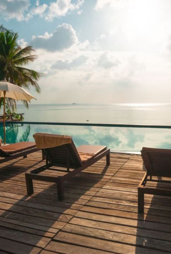 A beachfront resort deck with sun loungers, umbrellas, and a swimming pool facing the calm ocean at sunset.