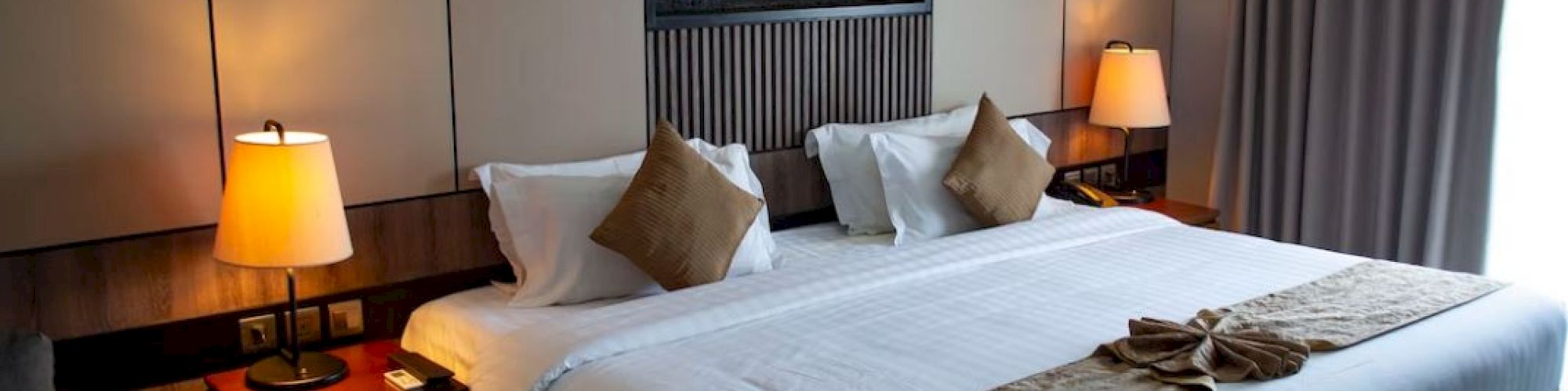 A modern hotel room with a neatly made bed, two bedside lamps, and a glowing fireplace feature in the wall.