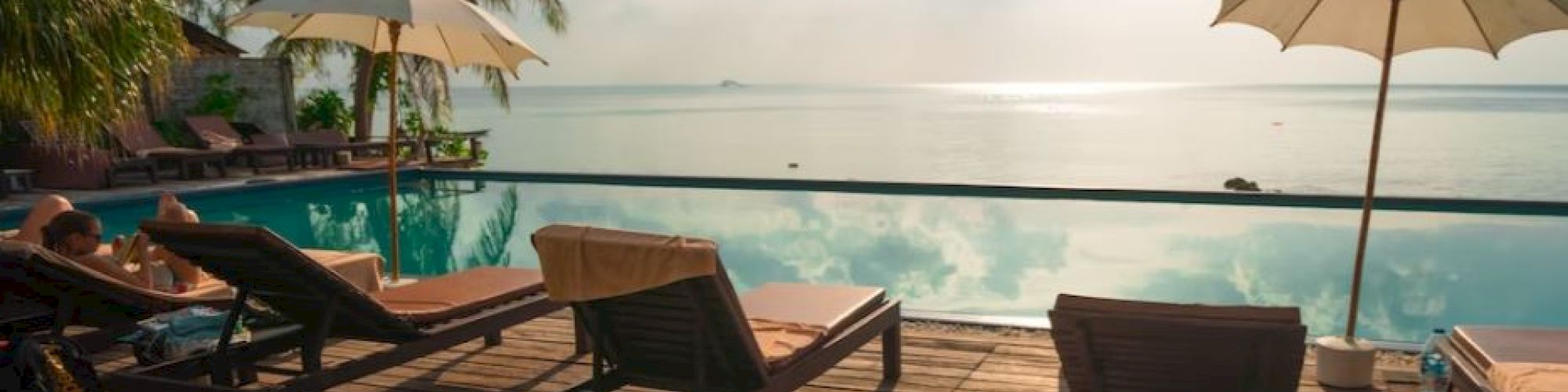 A tropical beach resort deck with sun loungers, umbrellas, palm trees, and calm ocean under a partly cloudy sky.