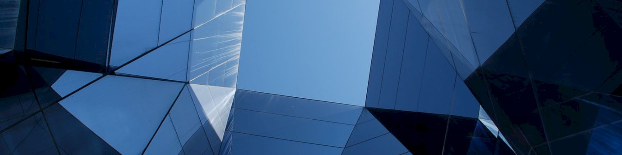 Geometric blue glass facade of a modern building, seen from below, with intersecting panels and a bright sky opening.
