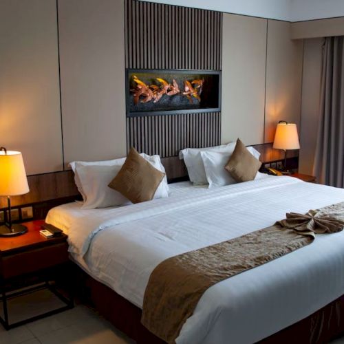 A cozy hotel room with a neatly made bed, bedside lamps, and a fireplace display of burning logs.