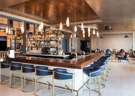 A modern hotel bar with a long wooden counter, lit bottles on shelves, hanging pendant lights, and stools for seating.