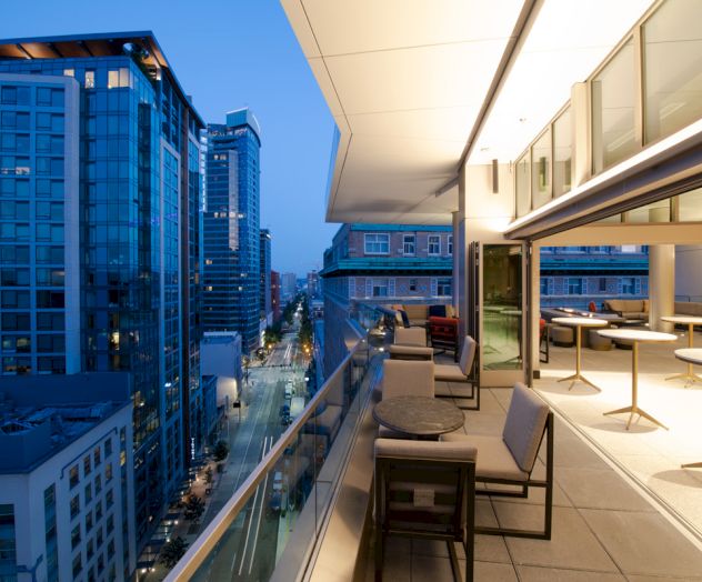 A modern high-rise rooftop terrace overlooks a busy city street between tall glass buildings at dusk.