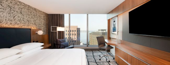 Modern hotel room with a large bed, desk chair, wall TVs, and a city view through big windows.