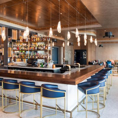 A modern hotel bar with a wooden counter, stocked bottle shelves, hanging pendant lights, and blue stools.