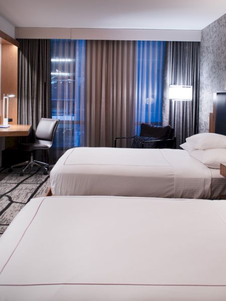 A modern hotel room with two beds, bedside lamps, a wall TV, desk area, and tall curtains covering the windows.