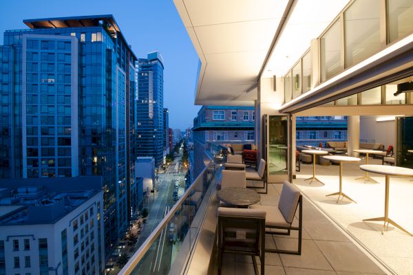 A high-rise balcony with lounge seating overlooks a city street and office towers under a twilight sky.