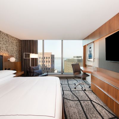 A modern hotel room with a large bed, desk chair, mounted TVs, wood paneling, and a view of city buildings through tall windows.