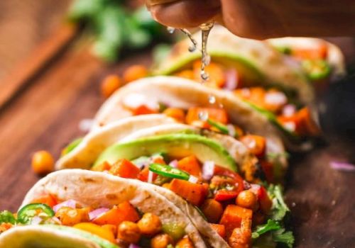 A person squeezes tacos with fresh veggie toppings and herbs, letting sauce drip over crunchy fillings.