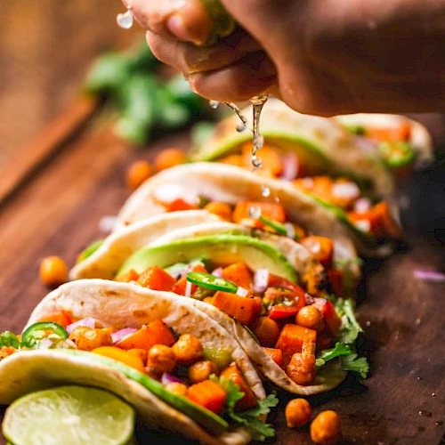 A person squeezes tacos with fresh veggie toppings and herbs, letting sauce drip over crunchy fillings.
