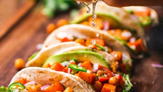 A person squeezes tacos with fresh veggie toppings and herbs, letting sauce drip over crunchy fillings.