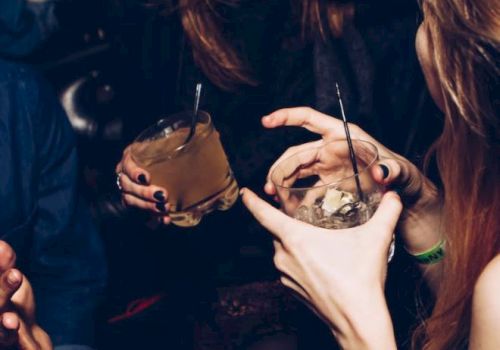 A party scene with two pierced people holding drinks with straws, smiling and posing together in a dim room.
