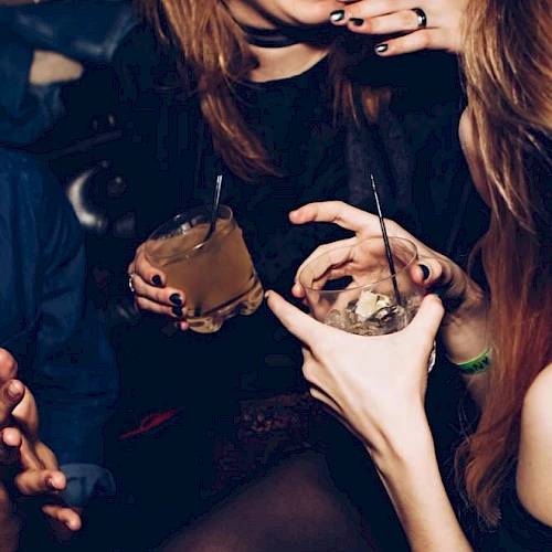 A party scene with two pierced people holding drinks with straws, smiling and posing together in a dim room.