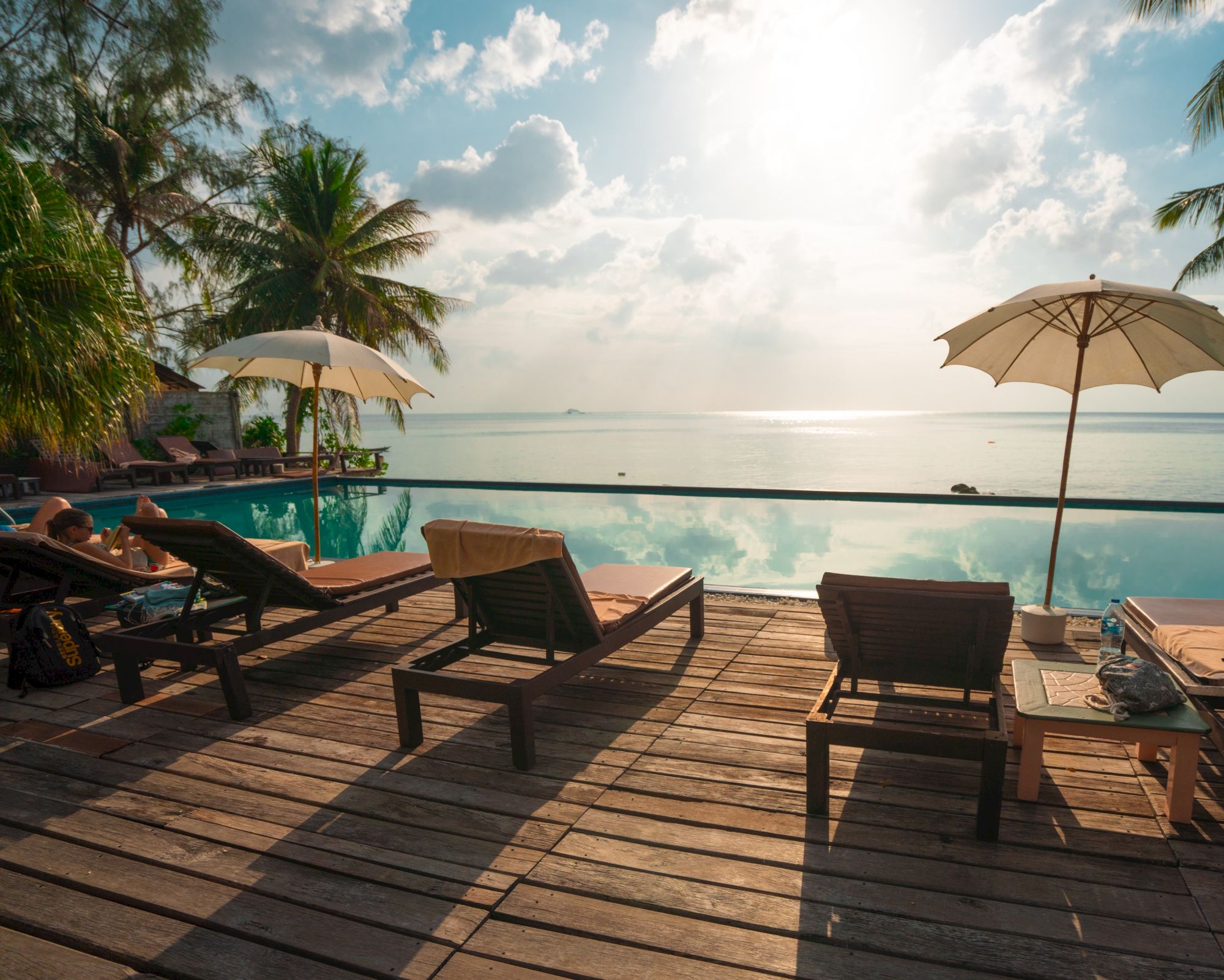 A tropical beachfront resort scene with a swimming pool, sun loungers, and umbrellas overlooking the ocean under a bright sky.