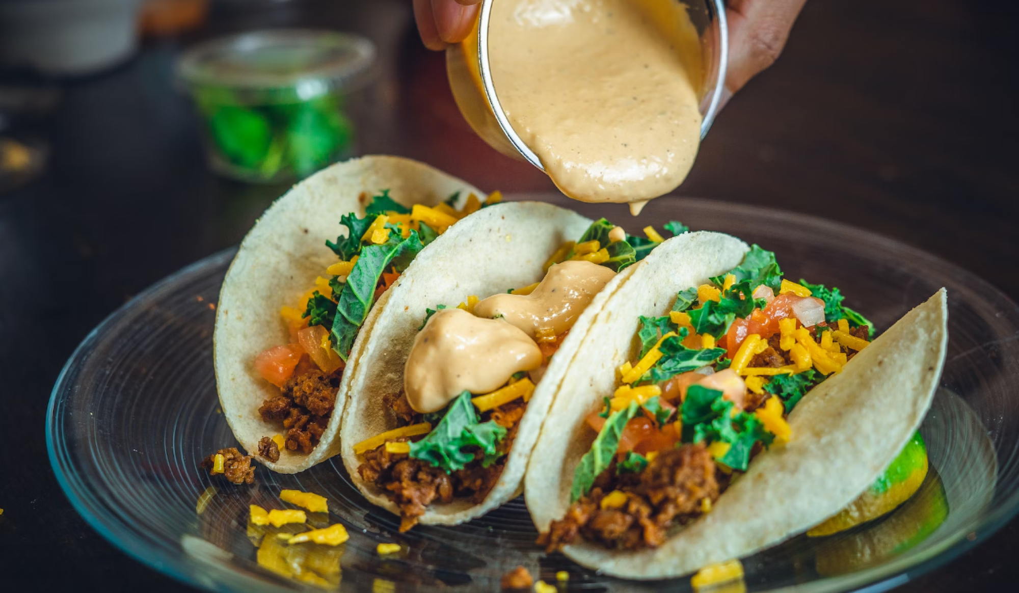 Three tacos on a plate with shredded lettuce, tomatoes, and cheese, with a creamy sauce being poured on top.