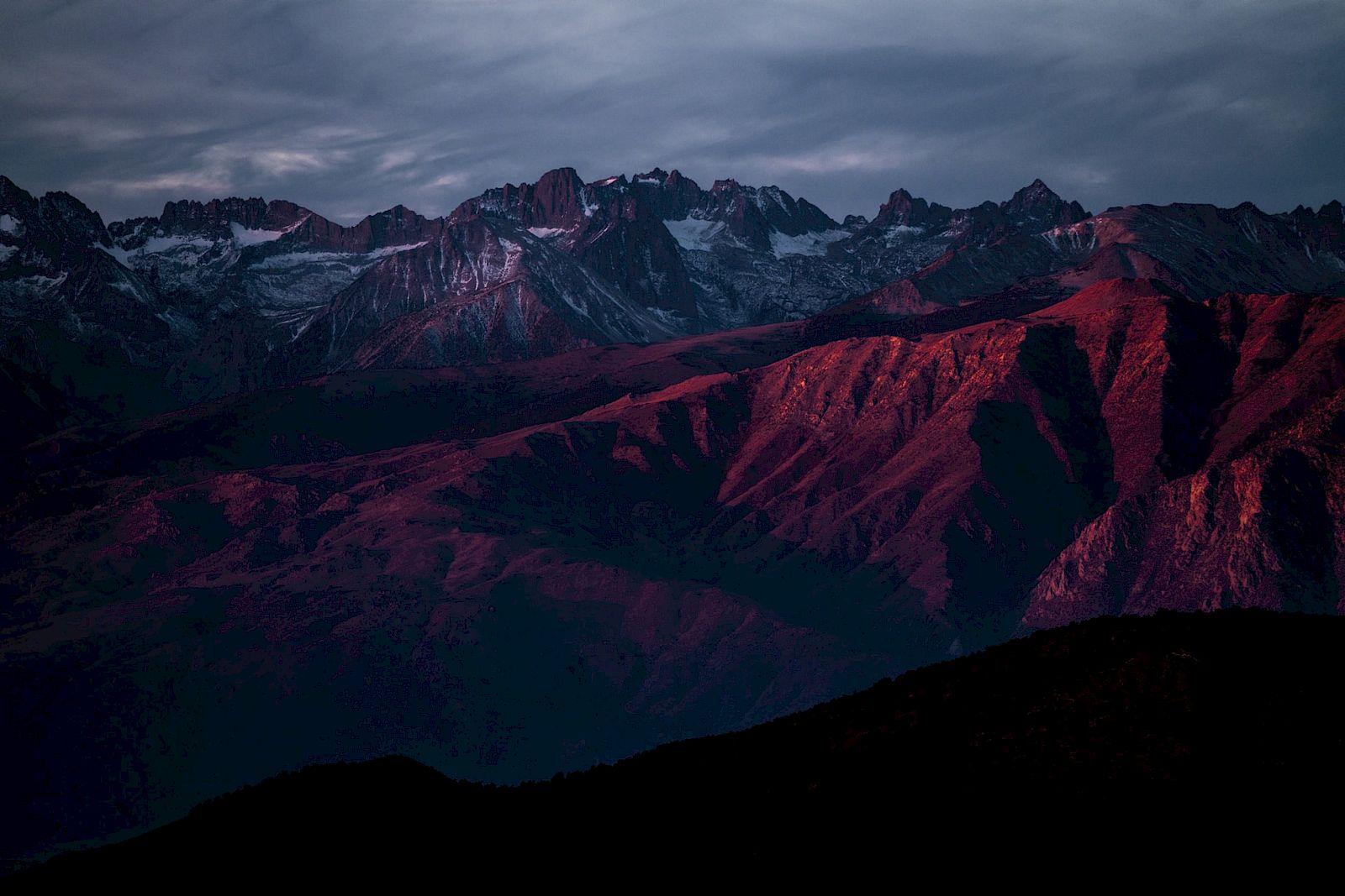 A dramatic mountain range at dusk, with snow-capped peaks and red-lit ridges under a cloudy sky.