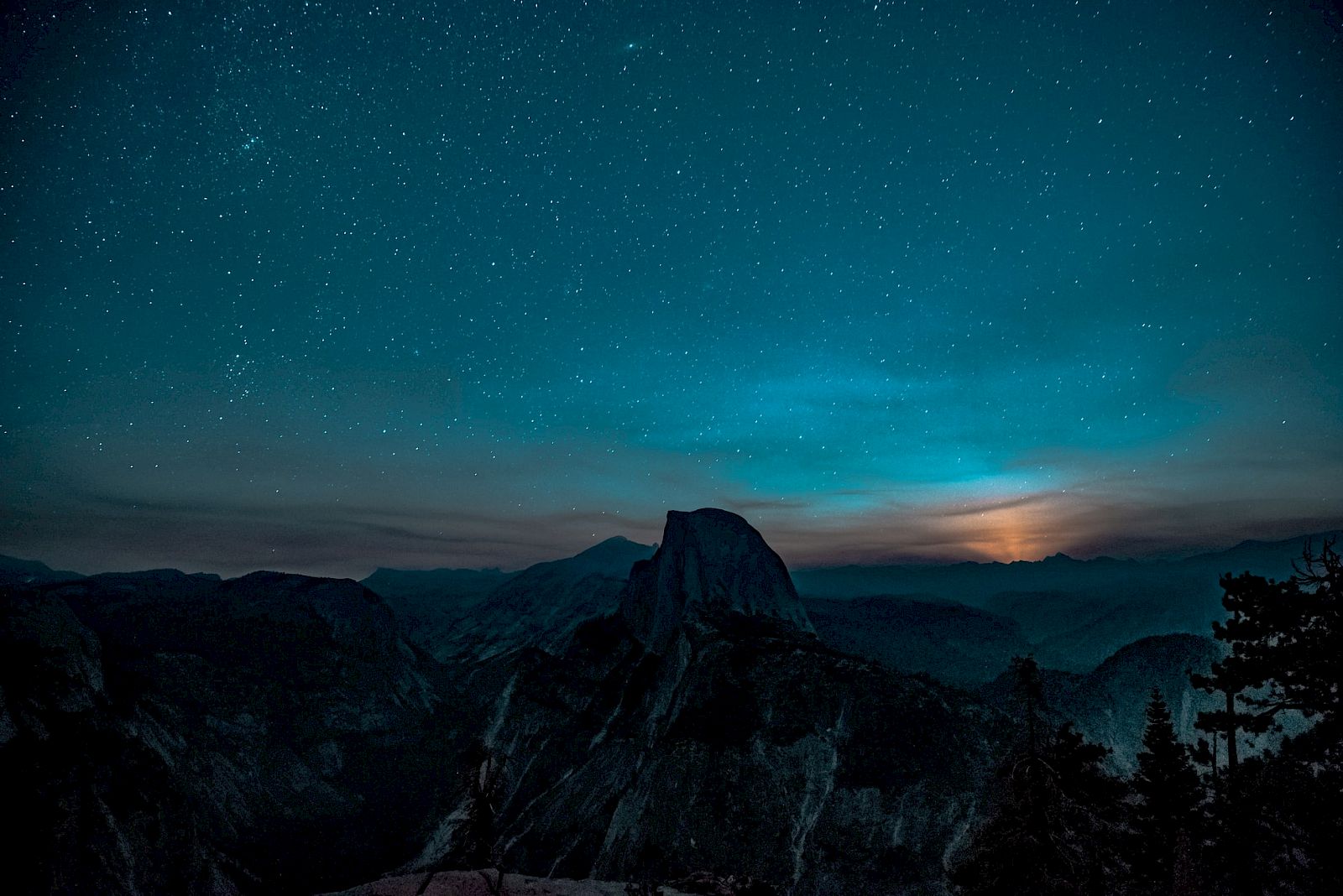 A star-filled night sky over dark mountain peaks, with a faint blue glow along the horizon.
