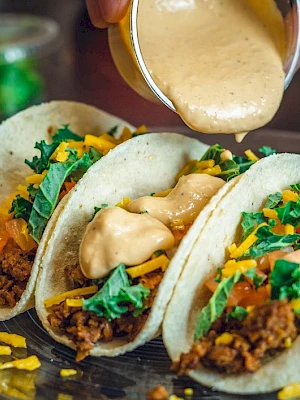 Three tacos on a plate with creamy sauce, shredded greens, cheese, and diced veggies, with more sauce being poured on top.