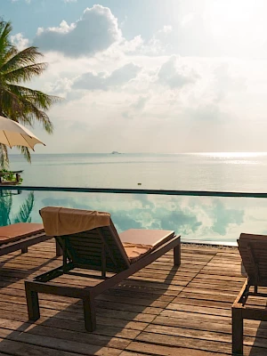A tropical resort deck with loungers under beige umbrellas, a pool, and palm views over a calm ocean.
