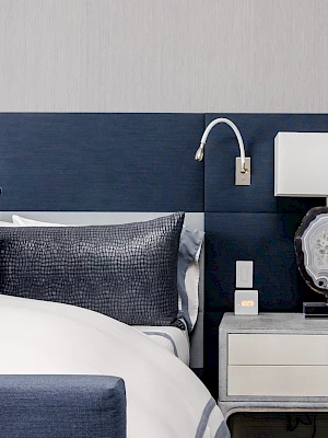 A modern bedroom with a neatly made bed, blue pillows, and a nightstand holding a decorative lamp and clock.