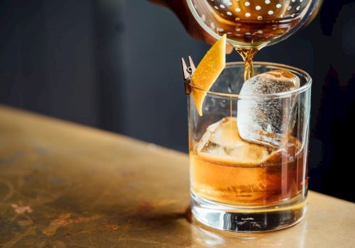 A whiskey or spirit on the rocks being poured into a glass with a citrus twist garnish, tall ice cube, and a drink being garnished.