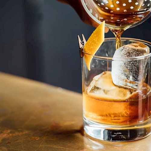 A whiskey or spirit on the rocks being poured into a glass with a citrus twist garnish, tall ice cube, and a drink being garnished.