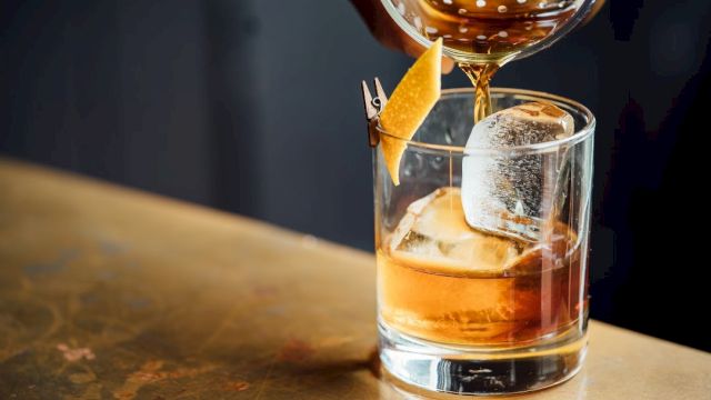 A whiskey or spirit on the rocks being poured into a glass with a citrus twist garnish, tall ice cube, and a drink being garnished.