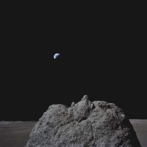 A tiny blue Earth floats above a dark rocky mound under a black night sky, like a surreal space scene.