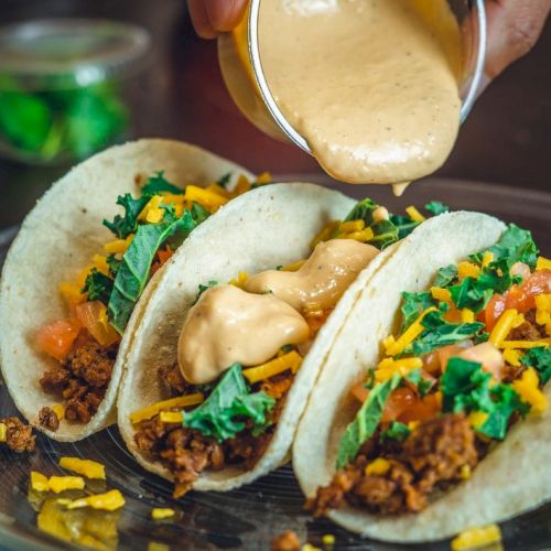 Three tacos on a plate as a creamy sauce is poured over them, topped with chopped greens and shredded cheese.