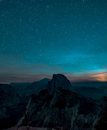 A star-filled night sky glows above dark mountain valleys, with misty clouds and a bright horizon light below.
