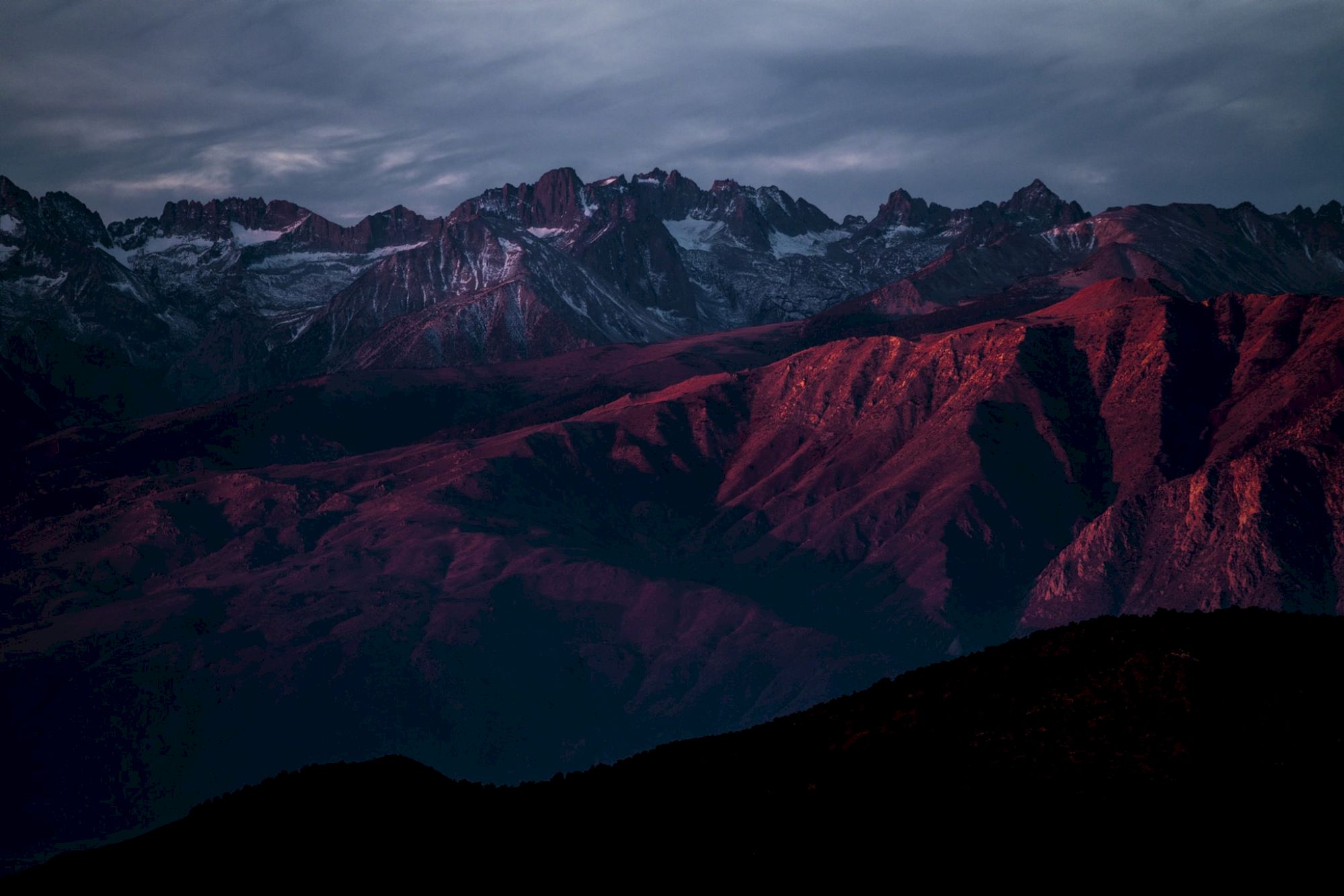 A dramatic mountain range at dusk with snowy peaks and crimson-lit ridges under a cloudy sky.