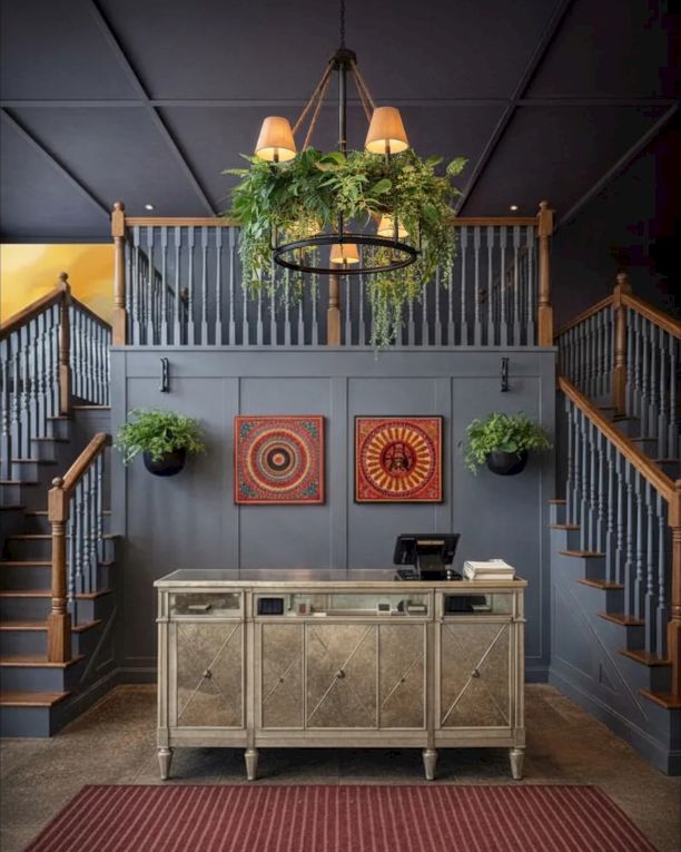 A stylish hotel lobby with a central metal credenza, two staircases, hanging greenery, and circular orange wall art beneath a black ceiling.
