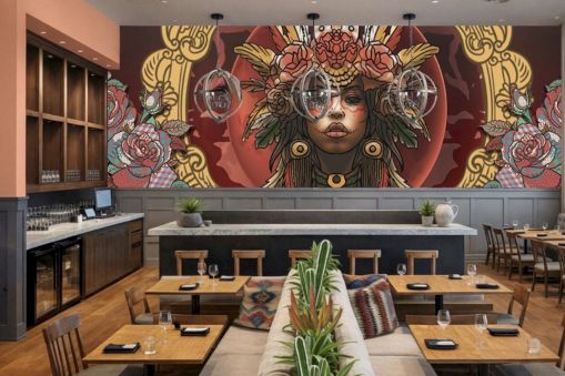 A stylish restaurant with a bold mural of a woman in ornate headdress on the back wall, wooden tables, plants, and modern decor.