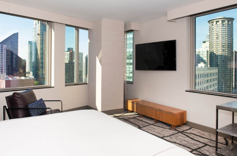 A sleek hotel room with a large bed, flat-screen TV, and city skyline view through big windows.