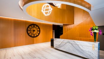 A modern office lobby with a marble reception desk, wood walls, neon globe light, wall clock, and a vase of purple flowers.