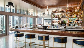 A sleek hotel bar with stools, a backlit liquor shelf, hanging lights, and a city skyline view through tall windows.