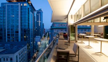 A modern high-rise rooftop balcony lounge overlooks a city street lined with tall buildings under a clear blue sky.