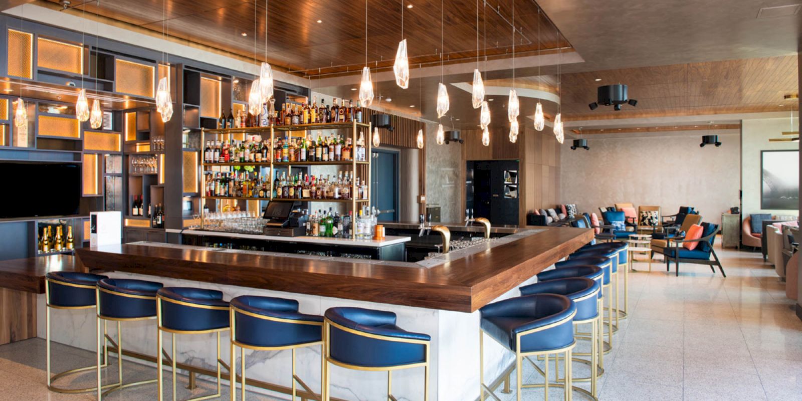 Modern bar lounge with a wood counter, bottle shelves, pendant lights, and blue chairs and stools.