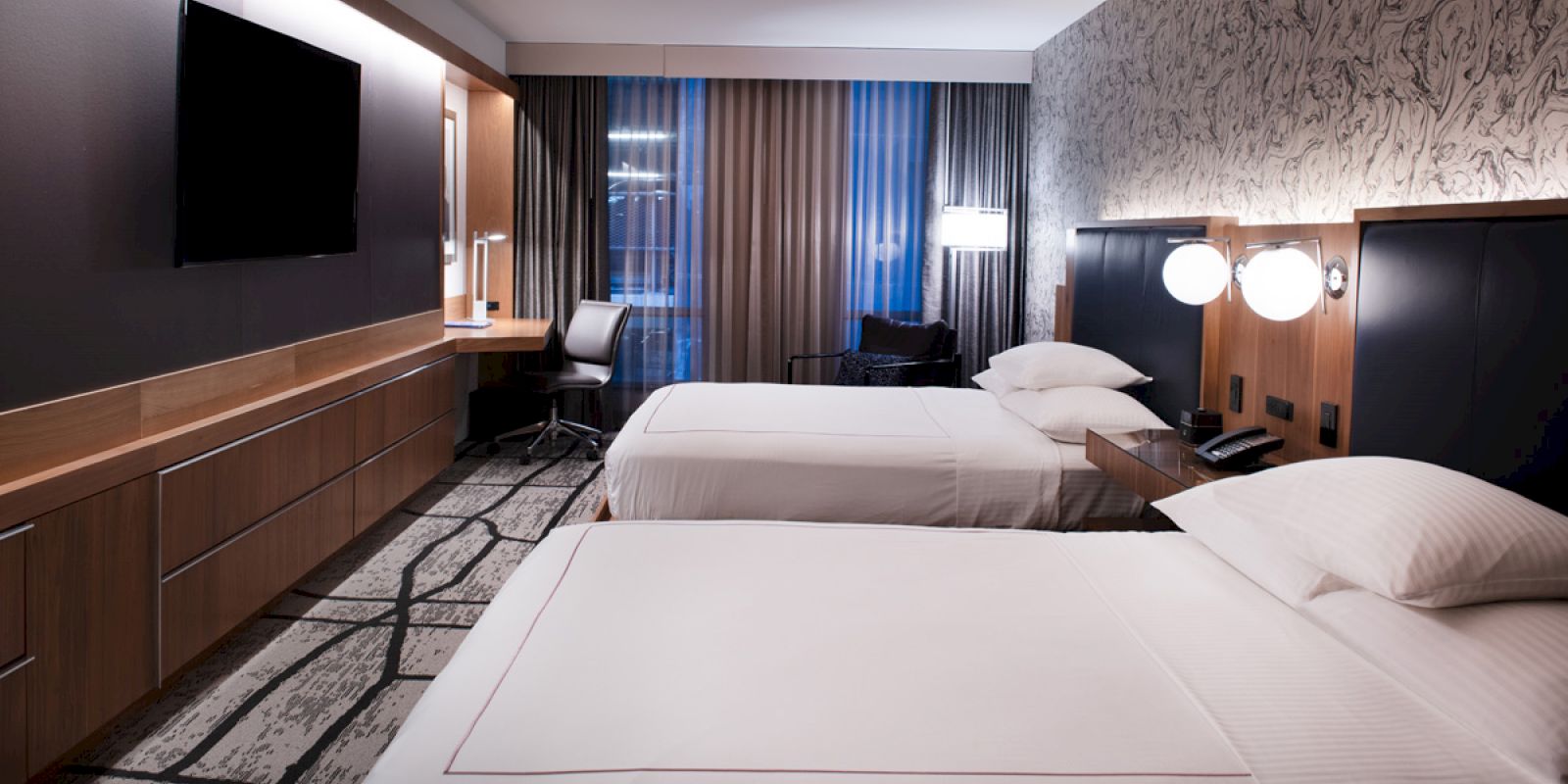 A modern hotel room with two white beds, black headboards, a flat-screen TV, and floor-to-ceiling curtains by the window.