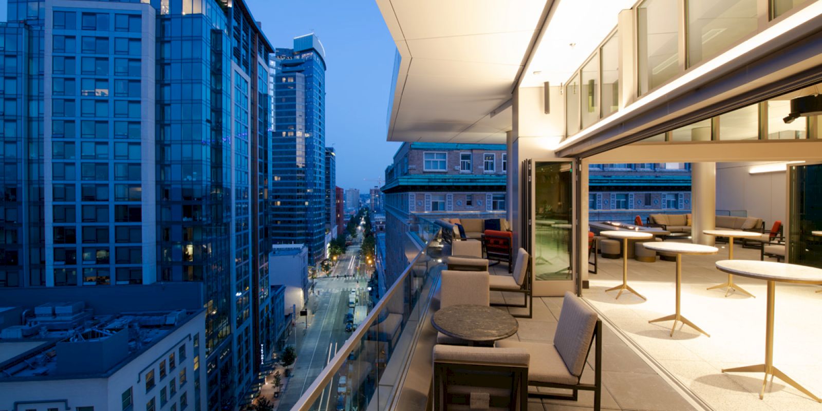 A modern high-rise rooftop terrace with lounge seating overlooks a downtown street and skyscrapers at dusk.