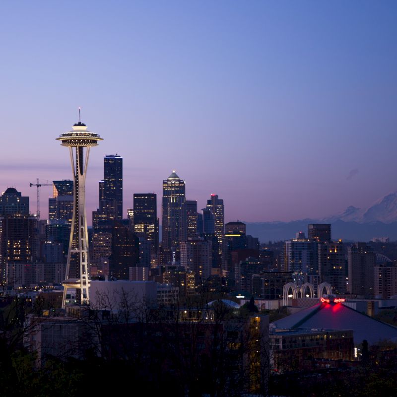 Seattle skyline at dusk with the Space Needle towering above city buildings and distant mountains, under a purple evening sky.