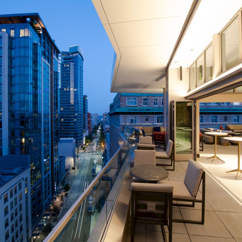 A rooftop terrace with modern lounge chairs overlooks a city street and tall office buildings under a blue evening sky.
