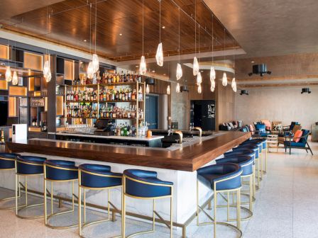 A modern hotel bar with a wood counter, bottle shelves, hanging lights, stools, and lounge seating.