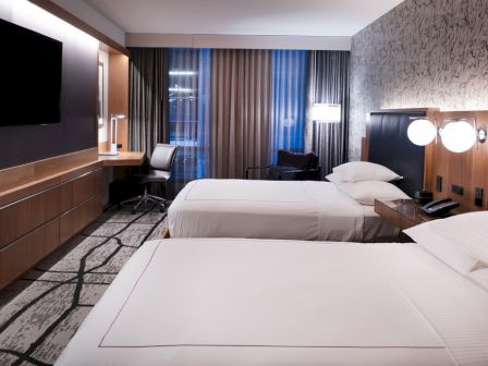 A modern hotel room with two neatly made beds, dark wood panels, curtains, a desk area, and floor-to-ceiling lighting.