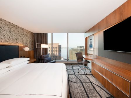 Modern hotel room with a bed, desk chair, large TVs, wood paneling, and big windows overlooking city buildings.