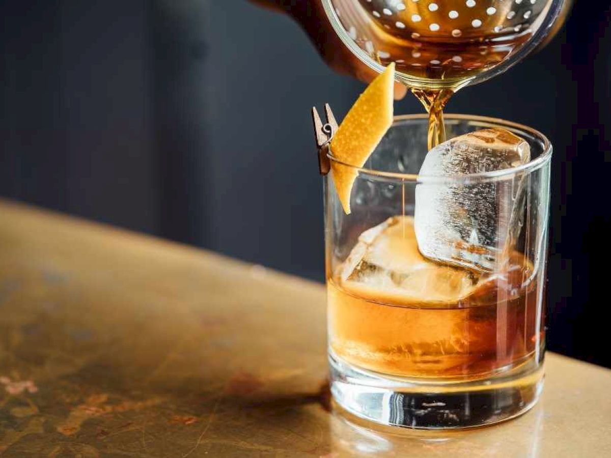 A martini glass pours an amber cocktail into a rocks glass with ice, garnished with a lemon twist.