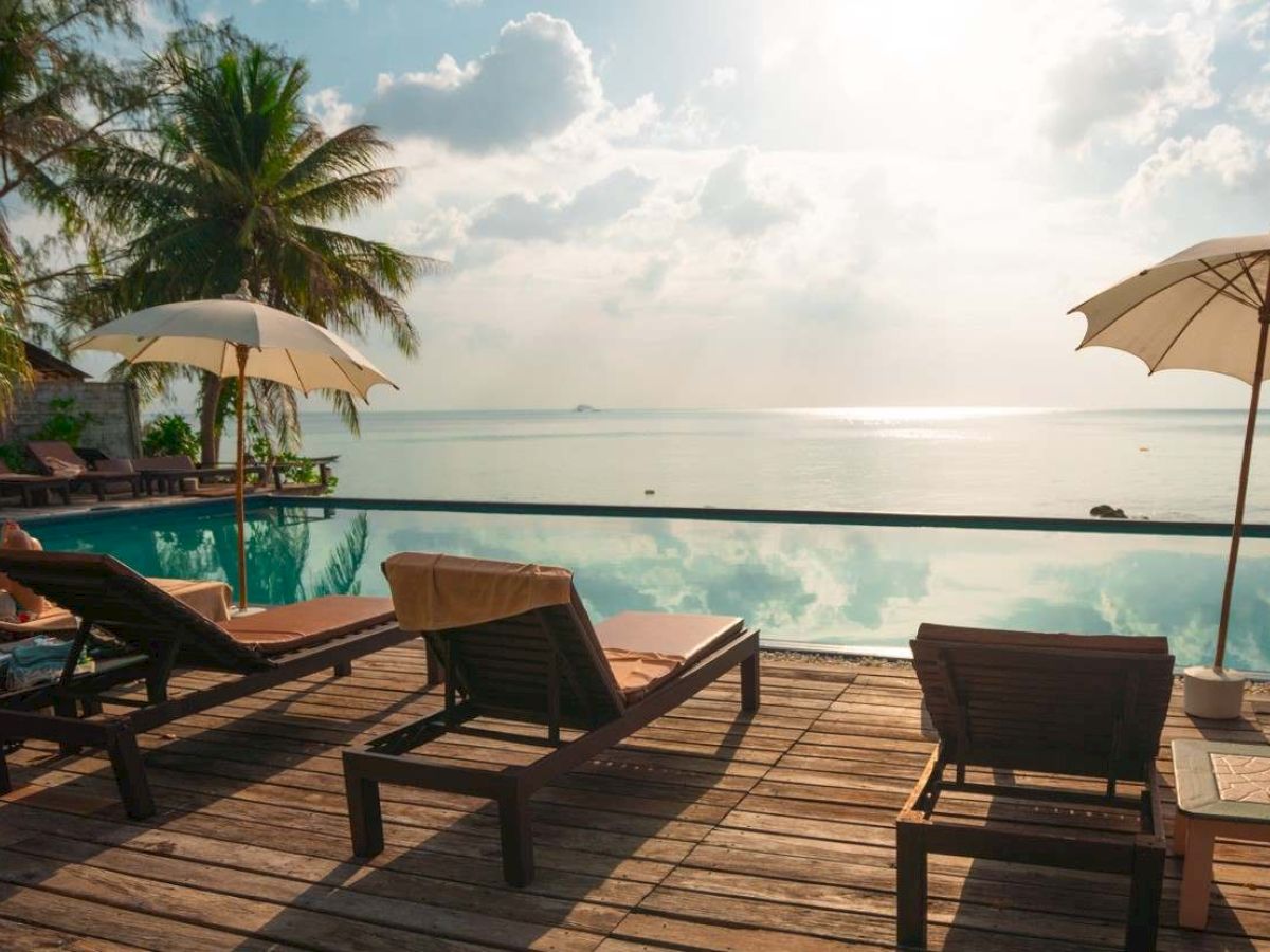Tropical beach resort scene with a pool, sun loungers, and umbrellas overlooking the ocean under a bright sky.